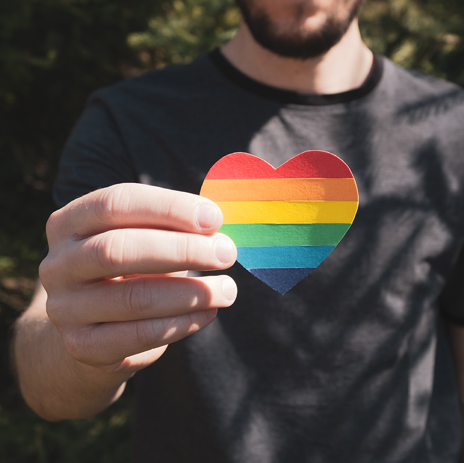 bearded young person holds up a heart made of rainbow-colored paper