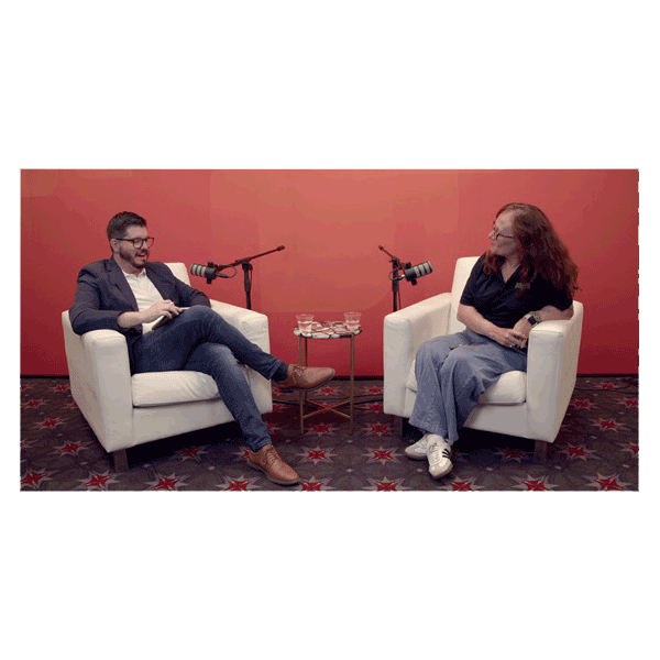a man and a woman sit in armchairs with microphones, speaking to each other